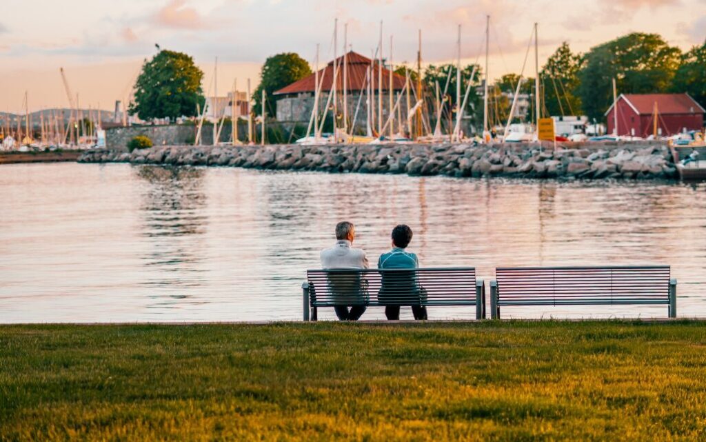 Par sitter på benk ved sjøen i Tresse, Kristiansand og ser utover båthavna og Festninga